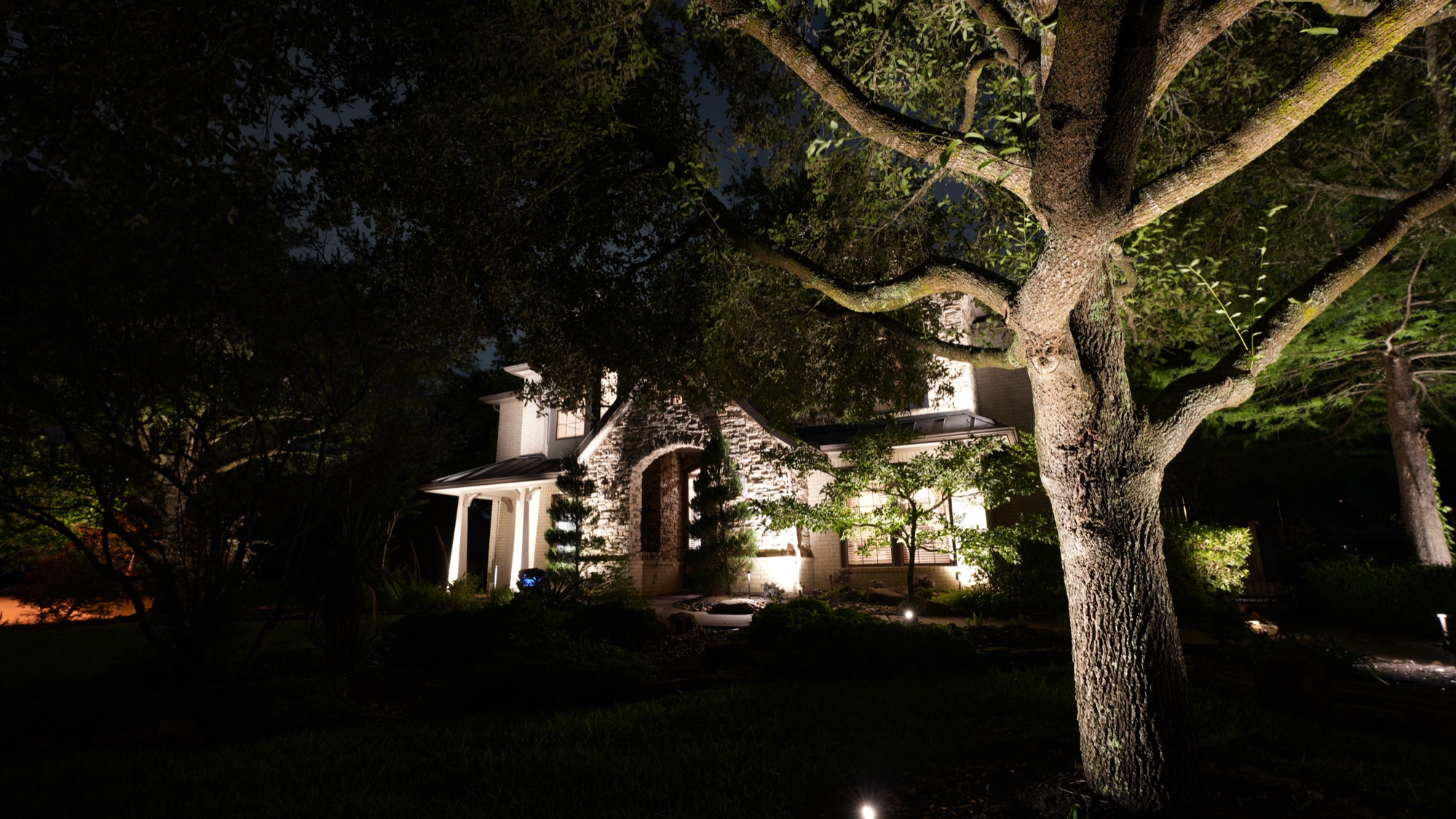 Landscape lighting on mature live oaks and stone house facade in Conroe, TX